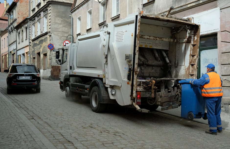 A street scene featuring a waste collection vehicle operated by a worker wearing a blue uniform and an orange high-visibility vest, with a matching blue cap. The vehicle is a large, white, front-loadi