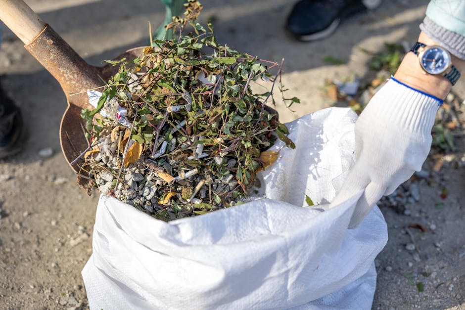 An aerial view of a patch of ground covered with a large accumulation of mixed waste, including discarded plastic bags, cardboard, and other debris, situated near a line of leafless trees and a fence.